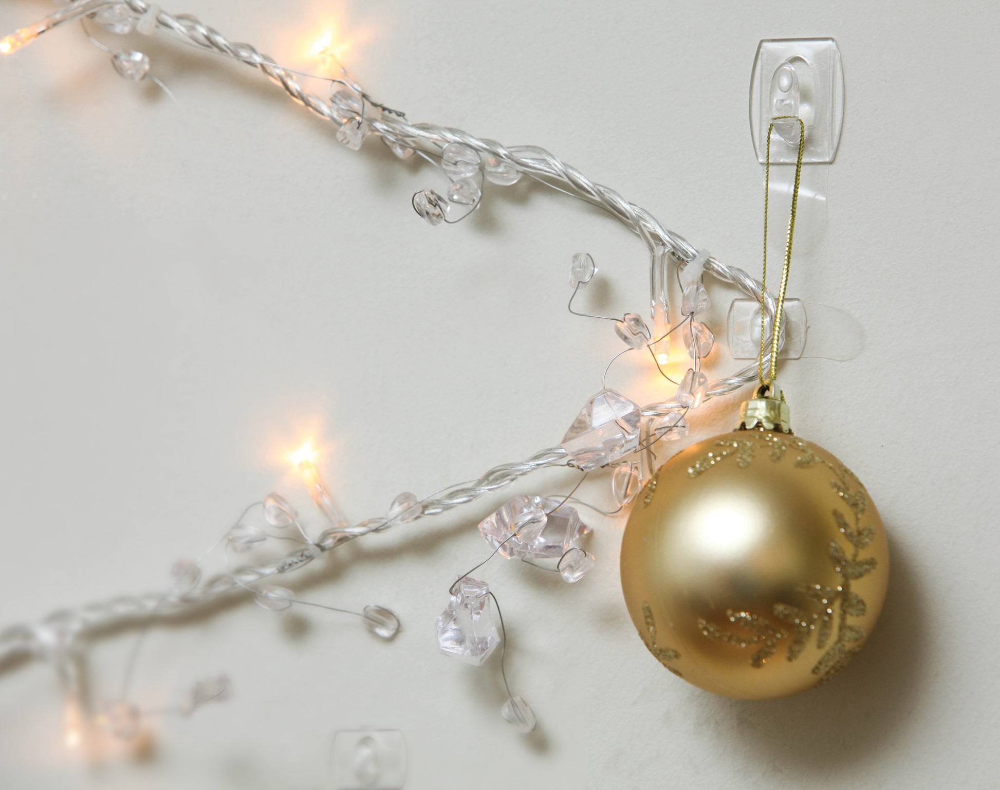 Ein goldener Weihnachtsschmuck hängt an einem Command™ Mini-Haken, transparent (3M Deutschland GmbH), der an einer weißen Wand neben weiß leuchtenden, mit Kunststoffkristallen verzierten Weihnachtslichtern angebracht ist.
