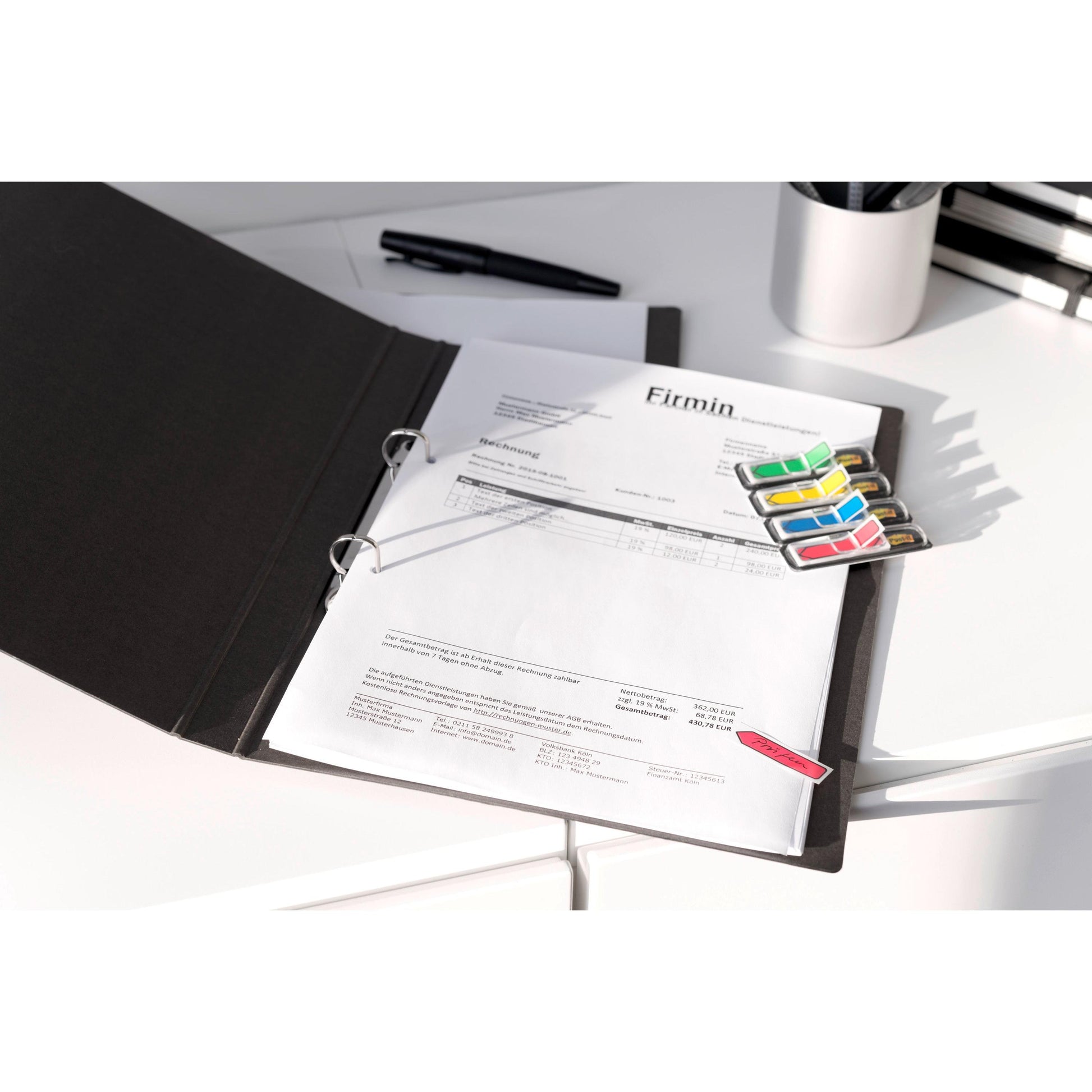Ein offener schwarzer Ordner mit gedruckten Dokumenten, bunten 3M Post-it® Index Pfeilen (11,9 mm x 43,2 mm, 24 Haftstreifen/Spender) und einem Stift auf einem weißen Schreibtisch; im Hintergrund sind Büroartikel der 3M Deutschland GmbH zu sehen.