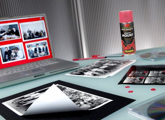 Auf einem Schreibtisch stehen Schwarz-Weiß-Fotos, ein Laptop mit Foto-Miniaturansichten, ein roter Stift, CDs, kleine rote Aufkleber und eine Dose 3M™ Scotch® PhotoMount™ Klebespray (400 ml) der 3M Deutschland GmbH auf einer hellen Fläche.