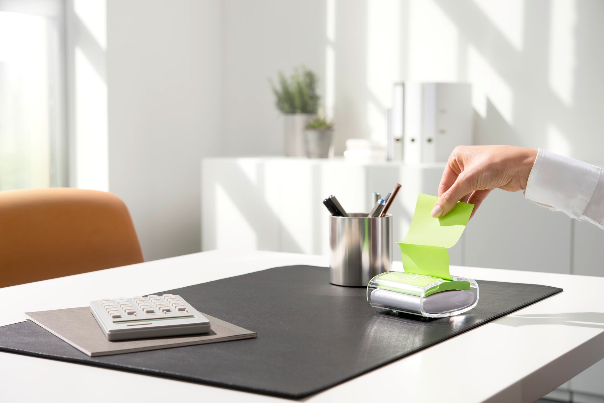 Eine Person zieht eine gelbe Post-it® Super Sticky Z-Note (76 x 76 mm) aus dem schwarzen PRO Post-it® Z-Notes Dispenser der 3M Deutschland GmbH, auf einem aufgeräumten Schreibtisch mit Taschenrechner, Stifthalter und schwarzer Unterlage bei hellem Tageslicht.
