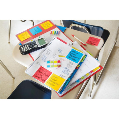 A student desk with notebooks, handwritten notes, a calculator, pens, highlighters, a folder, binders, papers, and Post-it® Super Sticky Notes (76 mm x 127 mm, 90 sheets/block) from 3M Deutschland GmbH, with empty chairs and a tiled floor in the background.