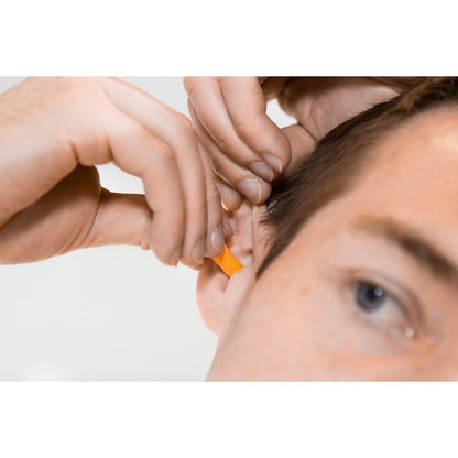 A person inserts an orange 3M™ earplug, without cord, two-pack, 1100P | 1 piece from 3M Deutschland GmbH into their ear with both hands. The image shows the ear, hands, and part of the face during careful application for noise reduction.