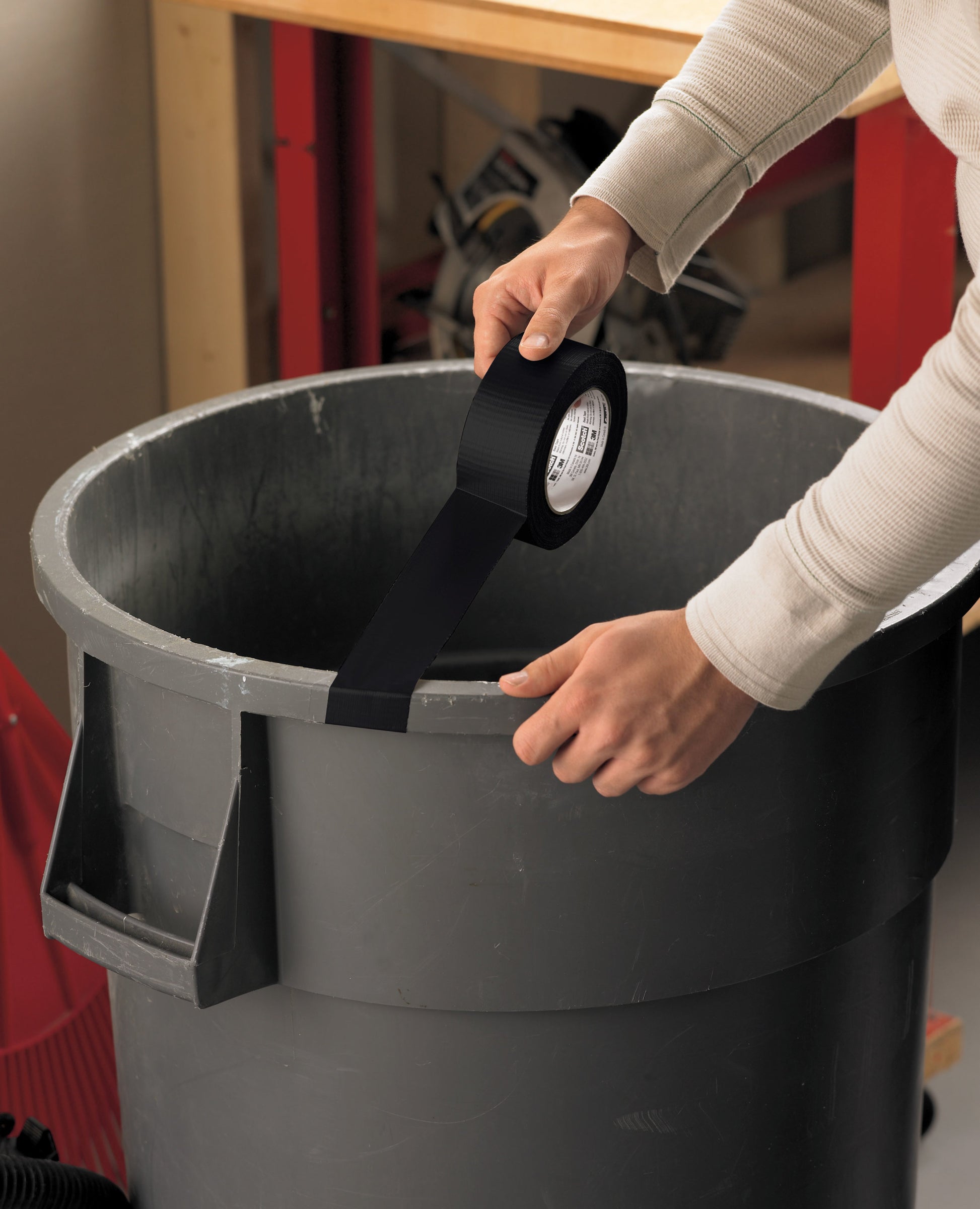 Eine Person klebt Scotch® Universal Klebeband 2904 (50 m x 48 mm) von 3M Deutschland GmbH auf den Rand eines großen grauen Abfalleimers in einer Werkstatt, mit einer Werkbank und Werkzeugen im Hintergrund.