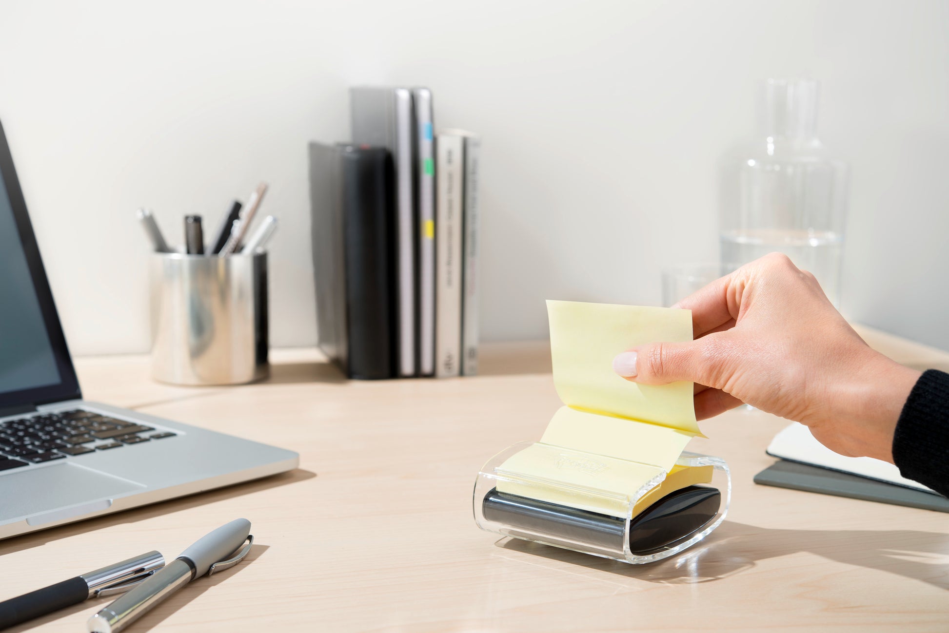 Eine Hand zieht eine gelbe Post-it® Super Sticky Z-Note (76x76 mm) aus dem schwarzen PRO Post-it® Z-Notes Dispenser der 3M Deutschland GmbH auf einem aufgeräumten Schreibtisch mit Laptop, Stiften, Büchern, Glaskaraffe und Tasse, die ordentlich im Hintergrund stehen.