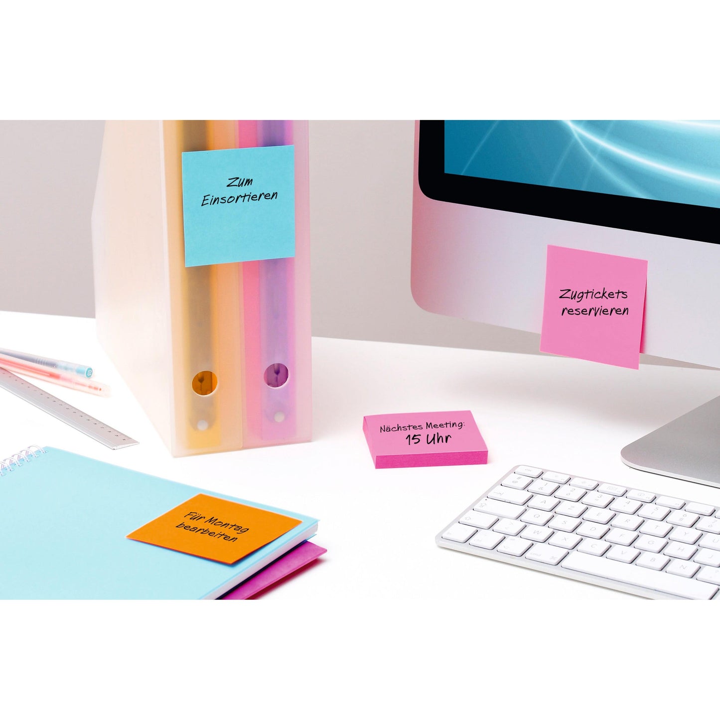 A desk with pastel-colored folders, a closed blue notebook, a keyboard, an iMac, and several 3M Deutschland GmbH Post-it® Super Sticky Notes Yellow (76 mm x 76 mm) with handwritten German reminders on folders, notebook, desk, and screen.