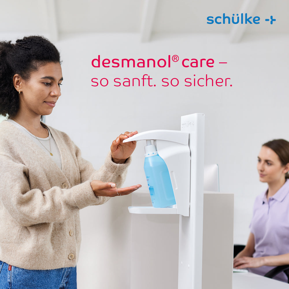 A woman uses a Schülke desmanol® care hand disinfectant dispenser at a counter while another woman sits nearby working on a computer. The text “desmanol® care – so gentle. so safe.” and the brand “Schülke & Mayr GmbH” are presented, with the skin care formula for gentle and effective hand disinfection being emphasized.